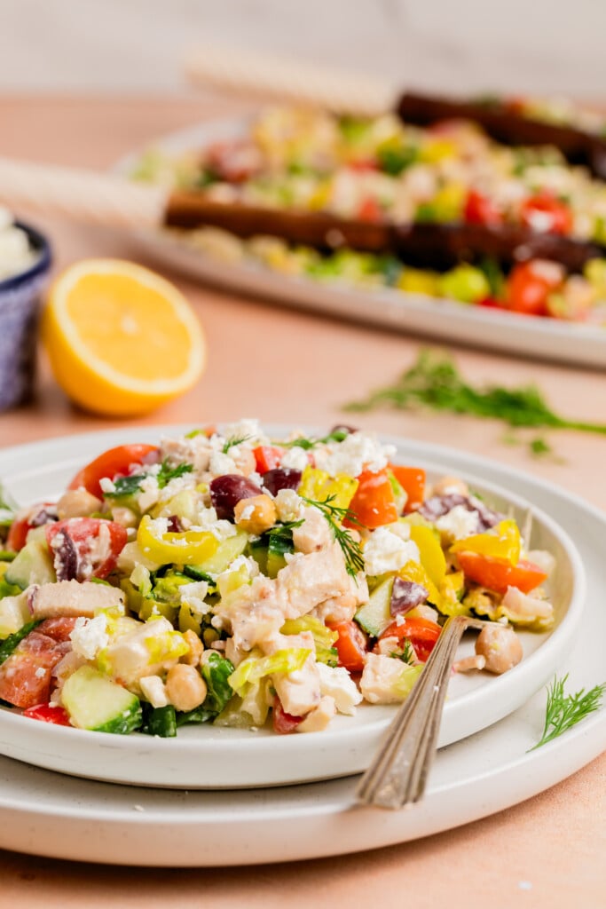 A colorful Mediterranean salad with chopped tomatoes, cucumbers, olives, chickpeas, peppers, and crumbled feta on a white plate, garnished with fresh herbs. A fork rests on the plate; lemon and a second plate are in the background.
