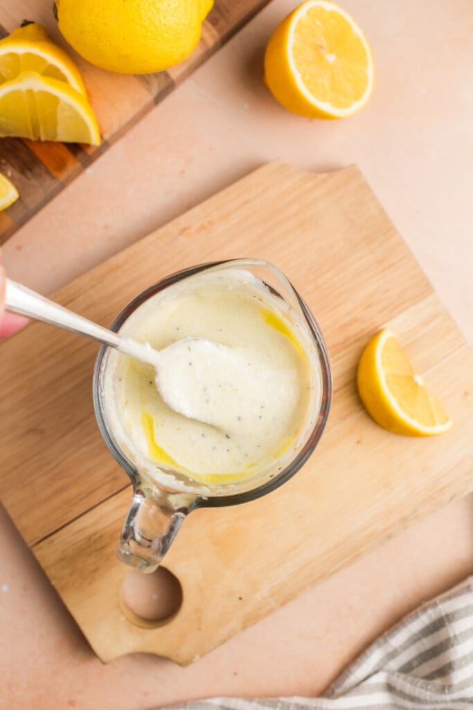 A glass measuring cup with creamy white dressing being stirred by a spoon, placed on a wooden cutting board next to lemon halves and slices. A striped cloth is partially visible in the corner.