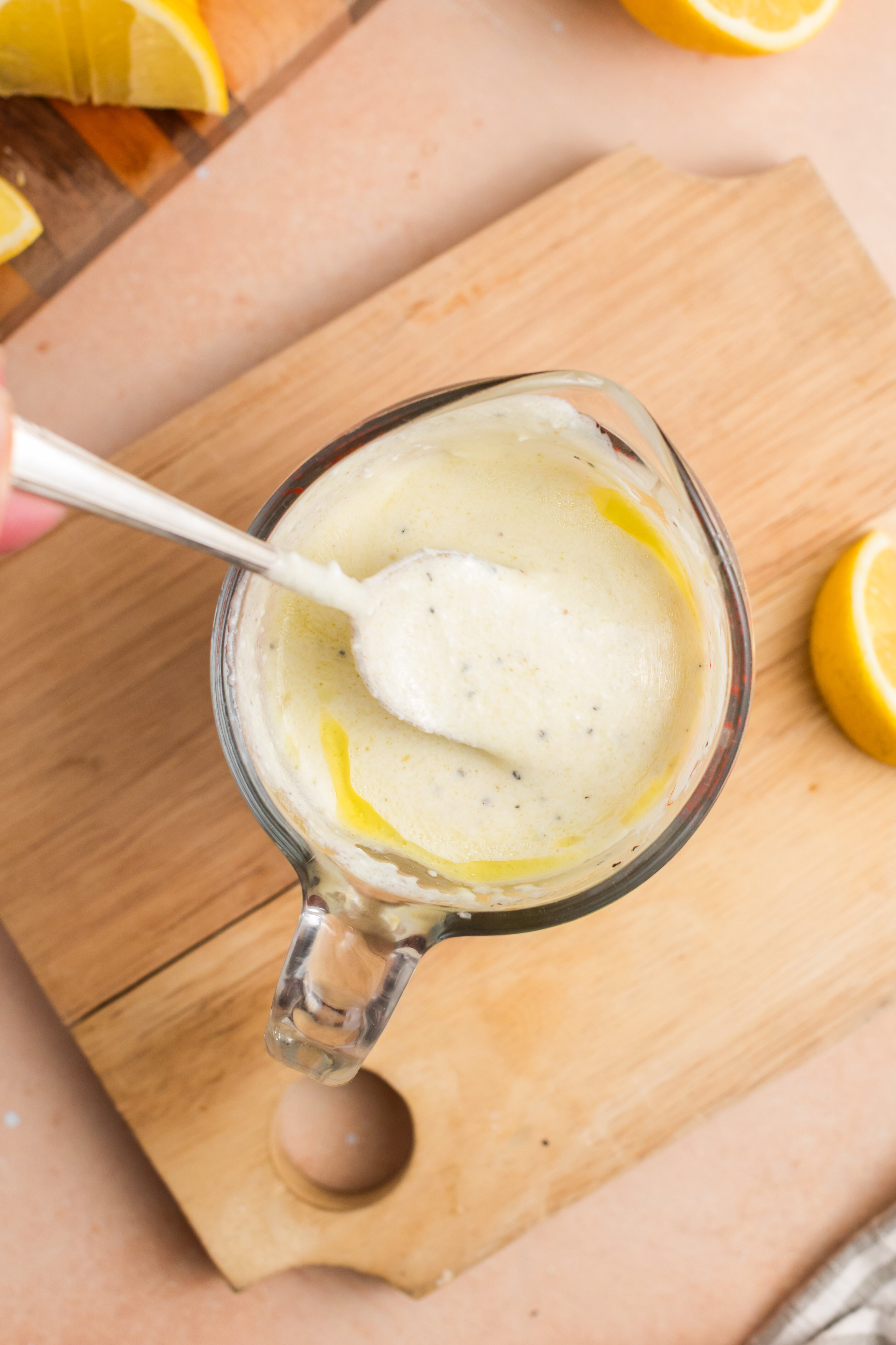 A glass measuring cup with creamy white dressing being stirred by a spoon, placed on a wooden cutting board next to lemon halves and slices. A striped cloth is partially visible in the corner.