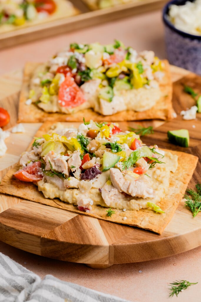 Two flatbreads topped with hummus, grilled chicken, diced tomatoes, cucumber, olives, lettuce, and fresh herbs are served on a wooden board. A bowl of creamy dip and salad ingredients are visible in the background.