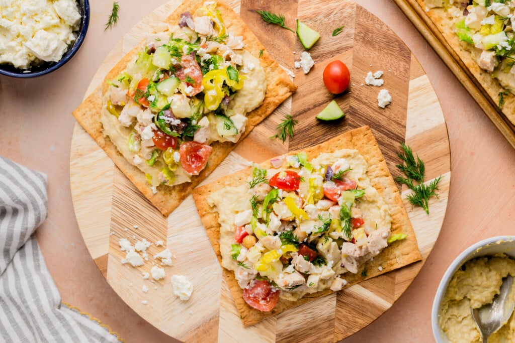 Rectangular flatbreads topped with hummus, diced chicken, tomatoes, cucumbers, peppers, and crumbled feta sit on a wooden board. Fresh herbs and small vegetable pieces are scattered nearby.