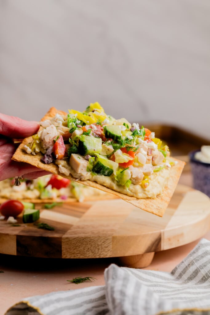 A hand holds a rectangular flatbread topped with diced vegetables, herbs, and what appears to be chicken over a wooden serving board. A striped cloth napkin sits in the foreground.