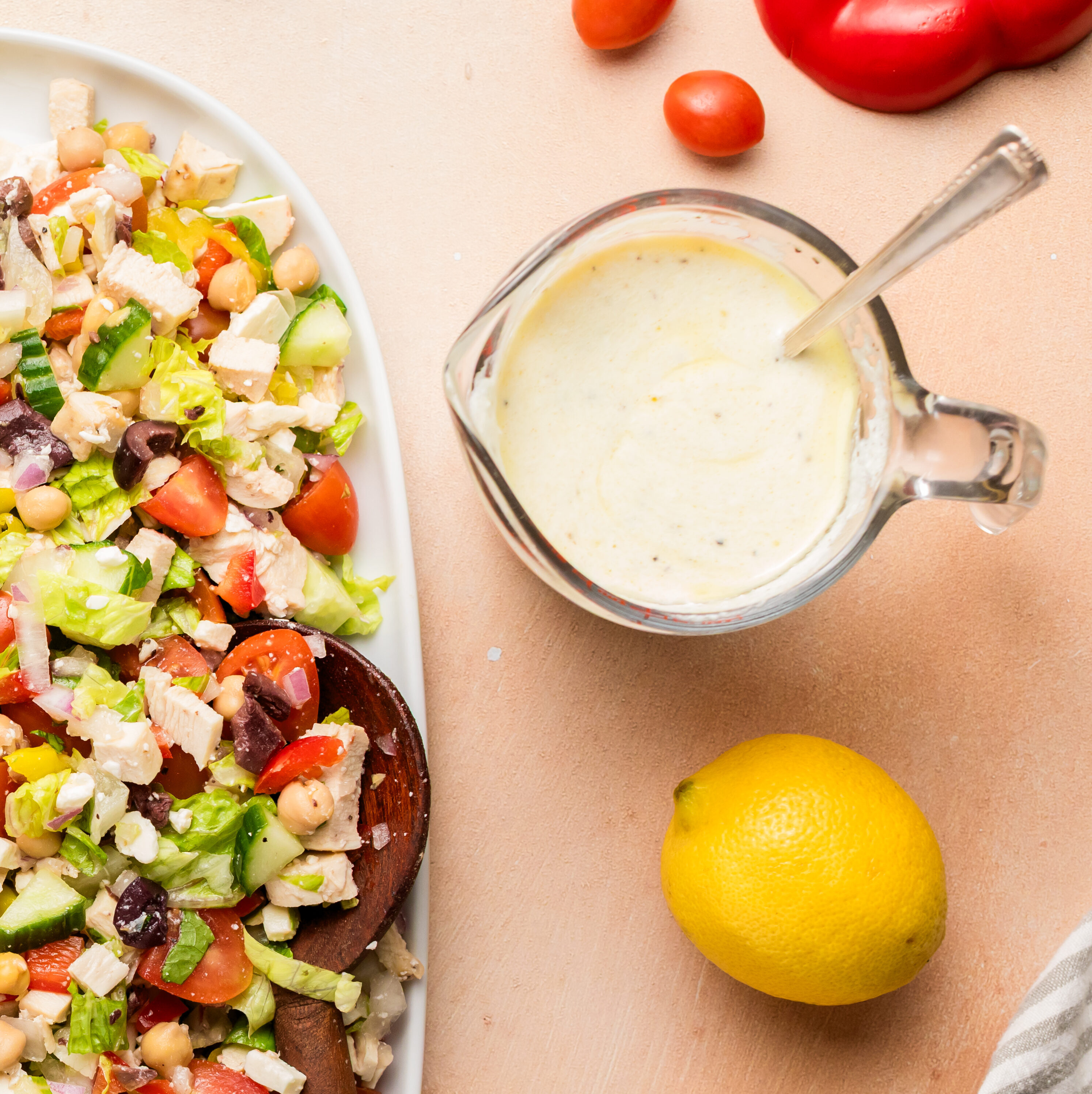 A large white platter holds a colorful chopped salad with tomatoes, cucumbers, olives, peppers, lettuce, and chicken, surrounded by lemons, feta cheese, salad dressing, and cut veggies on a peach-colored surface.