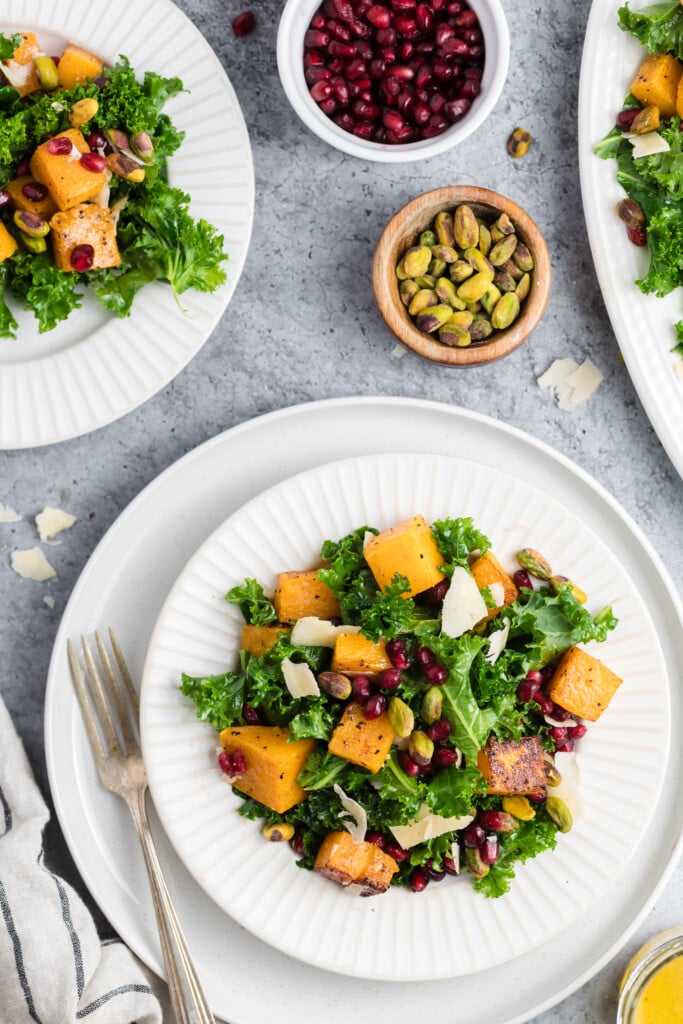 A white plate with kale salad topped with roasted squash, pomegranate seeds, pistachios, and cheese shavings, surrounded by bowls of pomegranate and pistachios on a gray surface. A fork rests beside the plate.