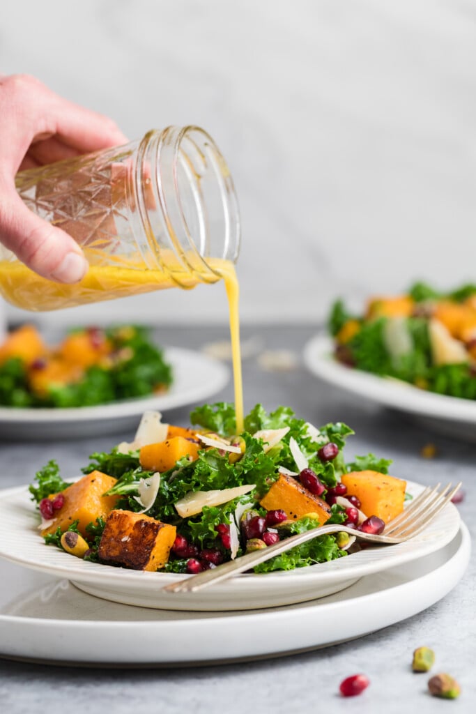A hand pours salad dressing from a jar onto a plate of kale salad with roasted butternut squash, pomegranate seeds, shaved cheese, and nuts. Other salad plates are blurred in the background.
