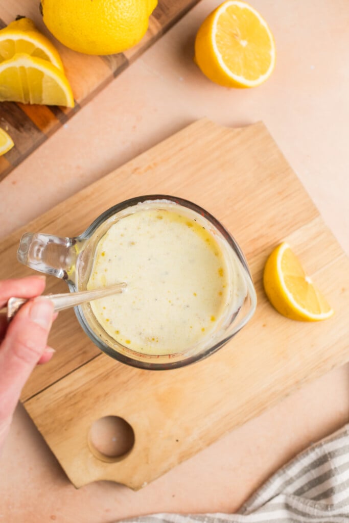 A hand stirs a creamy, yellow lemon vinaigrette in a glass measuring cup on a wooden cutting board, surrounded by sliced and halved lemons.