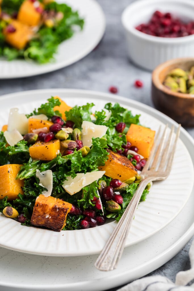 A fresh salad with kale, roasted butternut squash, pomegranate seeds, pistachios, and shaved cheese is served on a white plate with a fork. Small bowls of extra toppings are in the background.