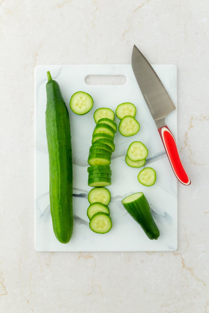 A whole cucumber, several cucumber slices, and a knife with a red handle are arranged on a white marble cutting board atop a light countertop.
