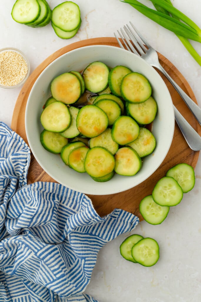 A bowl of sliced cucumbers on a wooden board, with extra cucumber slices, green onions, and sesame seeds nearby. A blue and white striped cloth and cutlery are also visible.