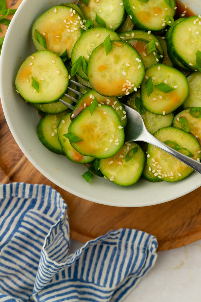 A bowl of sliced cucumbers garnished with sesame seeds and chopped green onions, with a fork resting inside. The bowl sits on a wooden board beside a blue and white striped cloth.