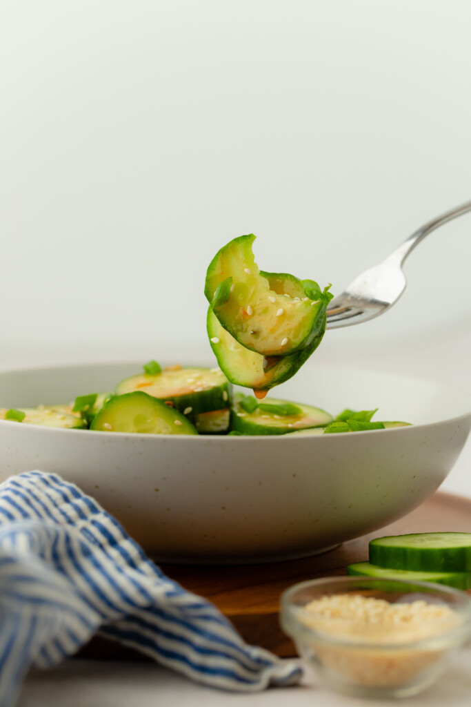A fork holds a bite of sliced cucumber above a bowl filled with cucumber salad. A blue and white striped cloth and a small dish of sesame seeds are visible nearby.