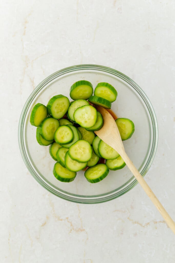 A glass bowl filled with sliced cucumbers and a wooden spoon, placed on a light-colored marble surface.