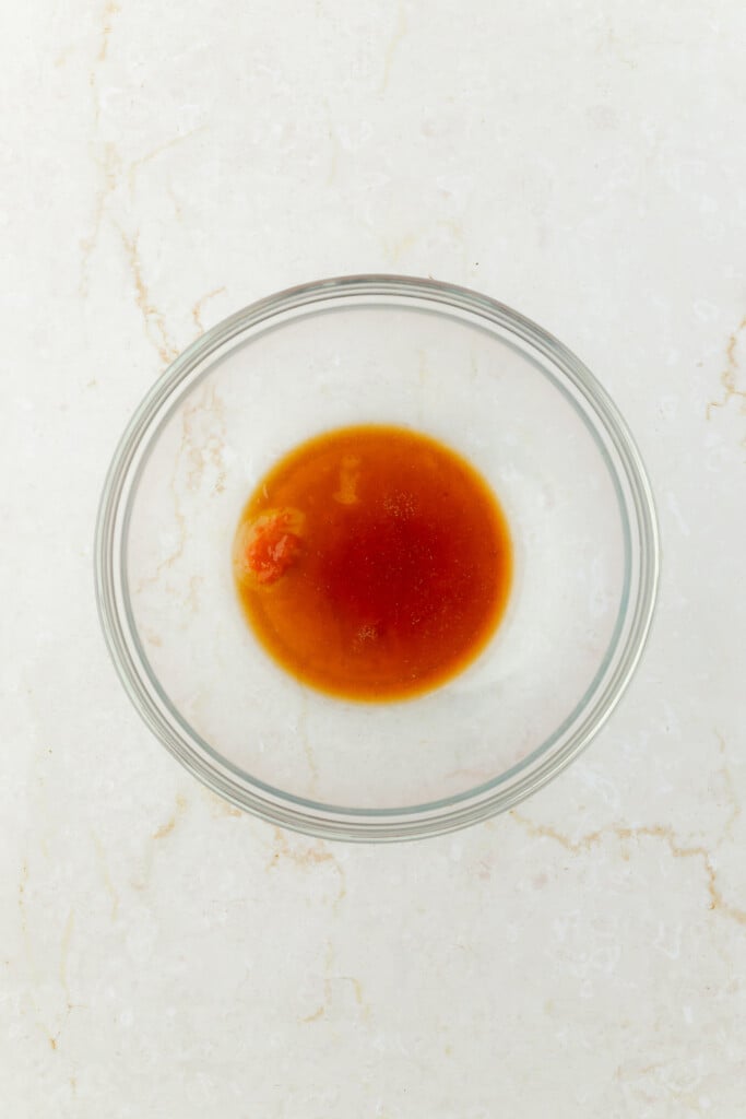 A small amount of reddish-brown liquid sauce in a clear glass bowl, placed on a light-colored marble surface.