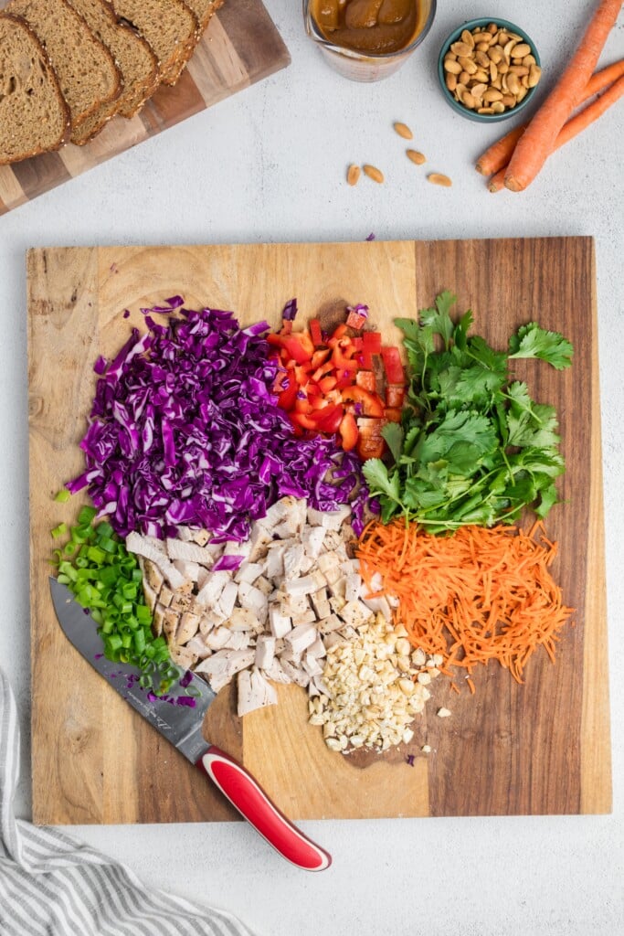 A wooden cutting board displays chopped ingredients: purple cabbage, red bell pepper, cilantro, shredded carrots, cooked chicken, green onions, and peanuts. A knife with a red handle rests nearby. Bread, carrots, and peanuts sit near the board.