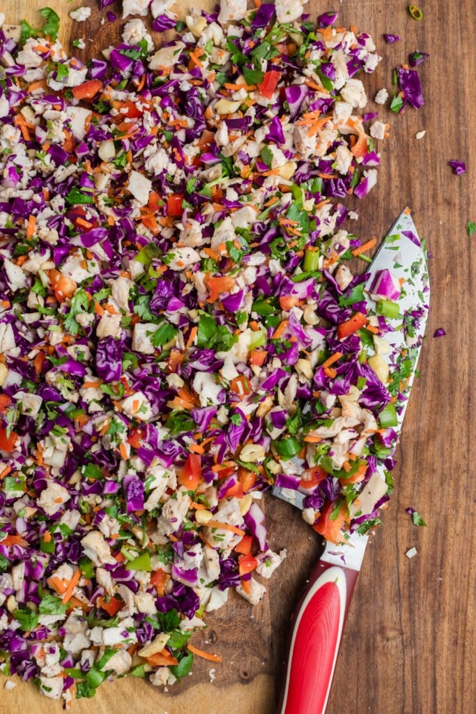 Chopped vegetables and herbs, including purple cabbage, red bell pepper, green onion, and parsley, spread on a wooden surface with a large chefs knife featuring a red handle.