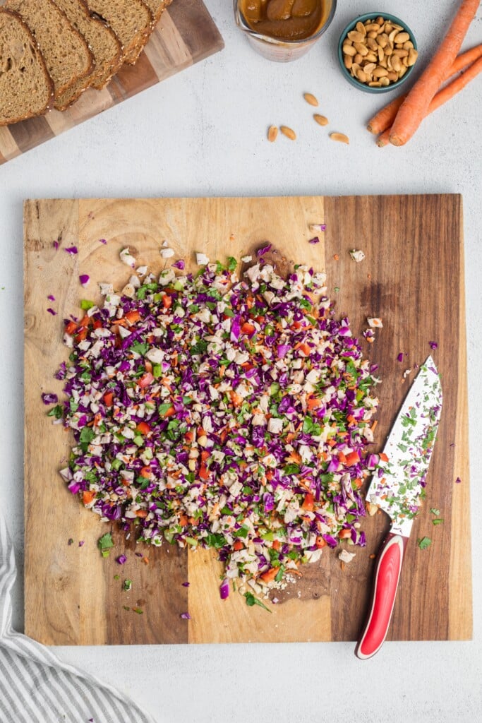 A wooden cutting board holds a colorful chopped salad with red cabbage, carrots, herbs, and peanuts. A floral-patterned knife with a red handle lies on the board. Sliced bread, peanuts, and carrots are nearby.