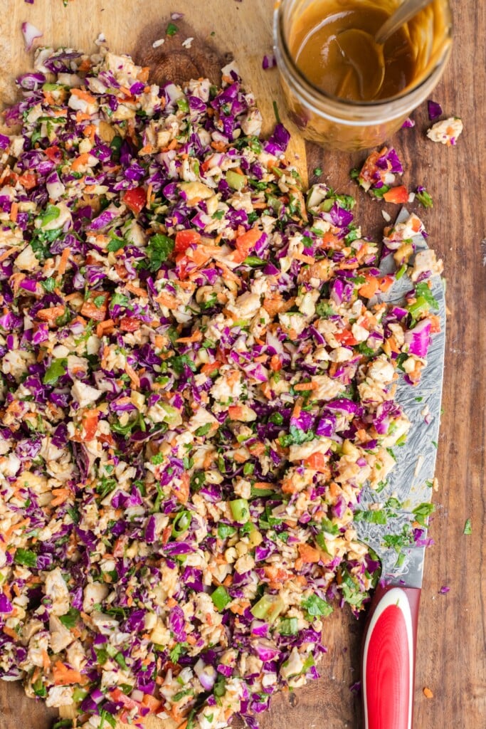 Chopped vegetable salad with red cabbage, carrots, green onions, and cilantro on a wooden cutting board next to a jar of brown dressing with a spoon. A knife with a red handle lies beside the salad.