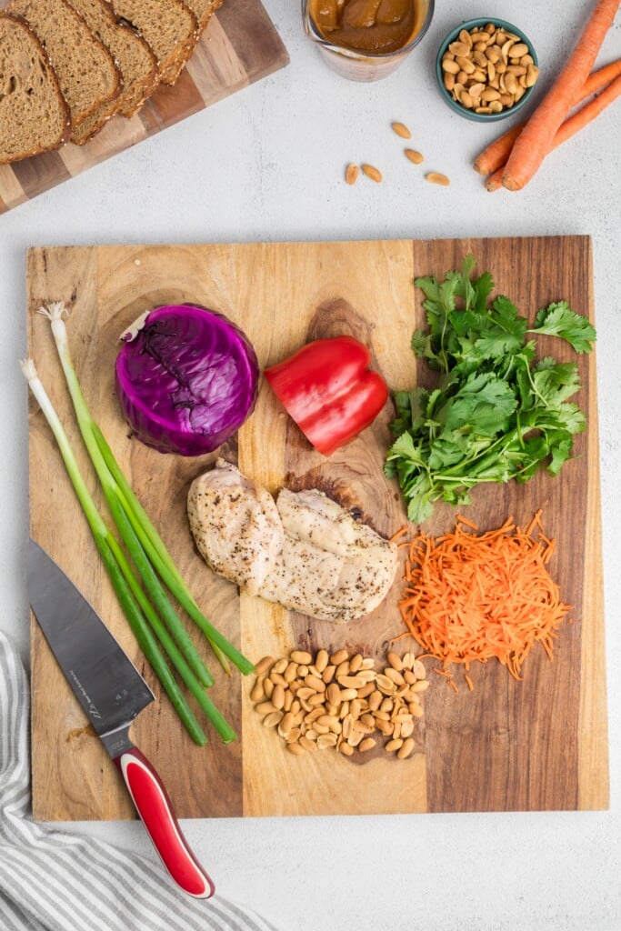 A wooden cutting board with cooked chicken breast, shredded carrot, red pepper, red cabbage, cilantro, green onions, and peanuts. Nearby are sliced bread, a can, a bowl of peanuts, carrots, and a knife with a red handle.