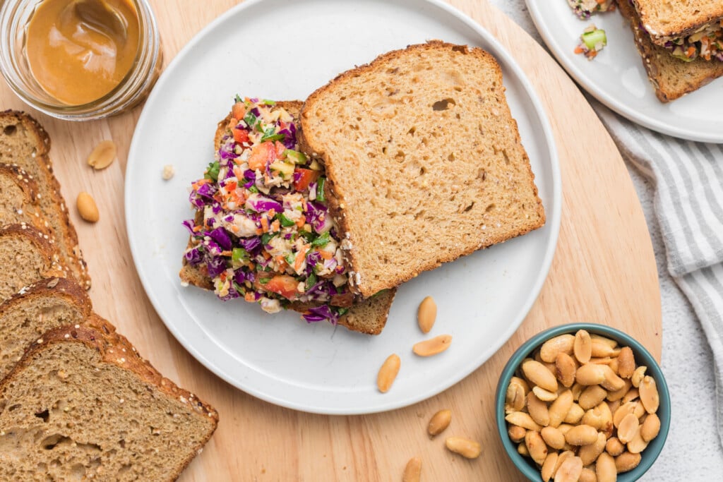 A sandwich with colorful vegetable filling on whole grain bread sits on a white plate, surrounded by peanuts, peanut butter, and extra bread slices on a wooden surface.