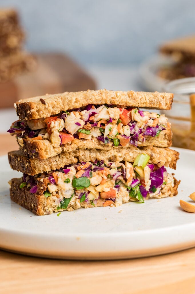 A stacked sandwich filled with colorful chopped vegetables and what appears to be tofu or tempeh, served on whole grain bread. The sandwich sits on a white plate with peanuts and a jar of sauce in the background.