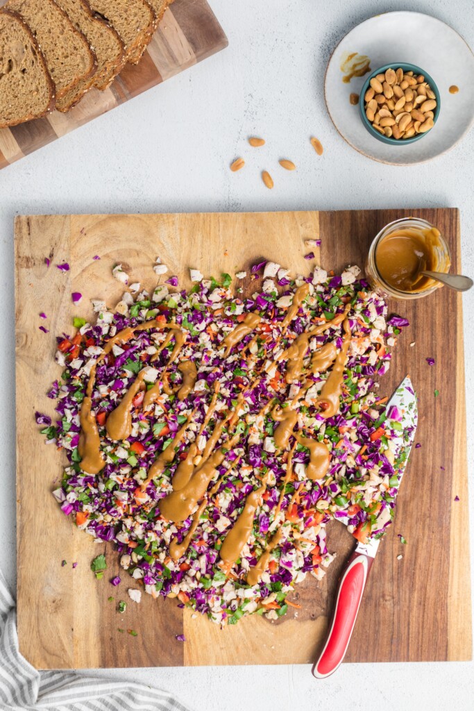 A wooden cutting board with chopped colorful vegetables topped with a drizzle of peanut sauce, a knife with a red handle, a bowl of peanuts, a small bowl of extra sauce, and slices of bread on the side.