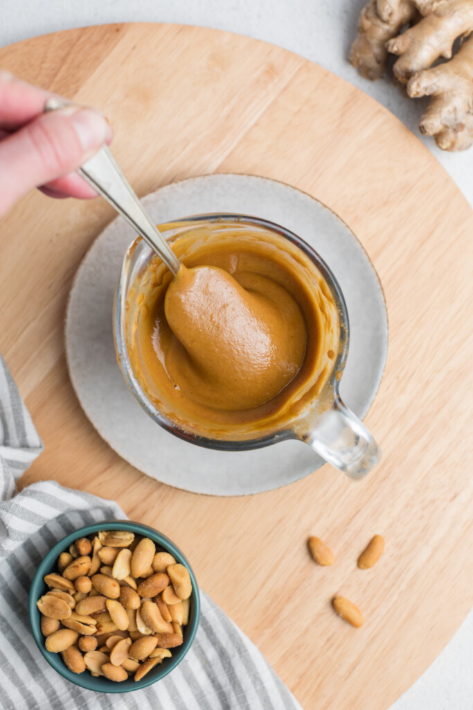 A hand stirring creamy peanut butter in a glass cup on a wooden board, with a small bowl of peanuts and a piece of ginger nearby.