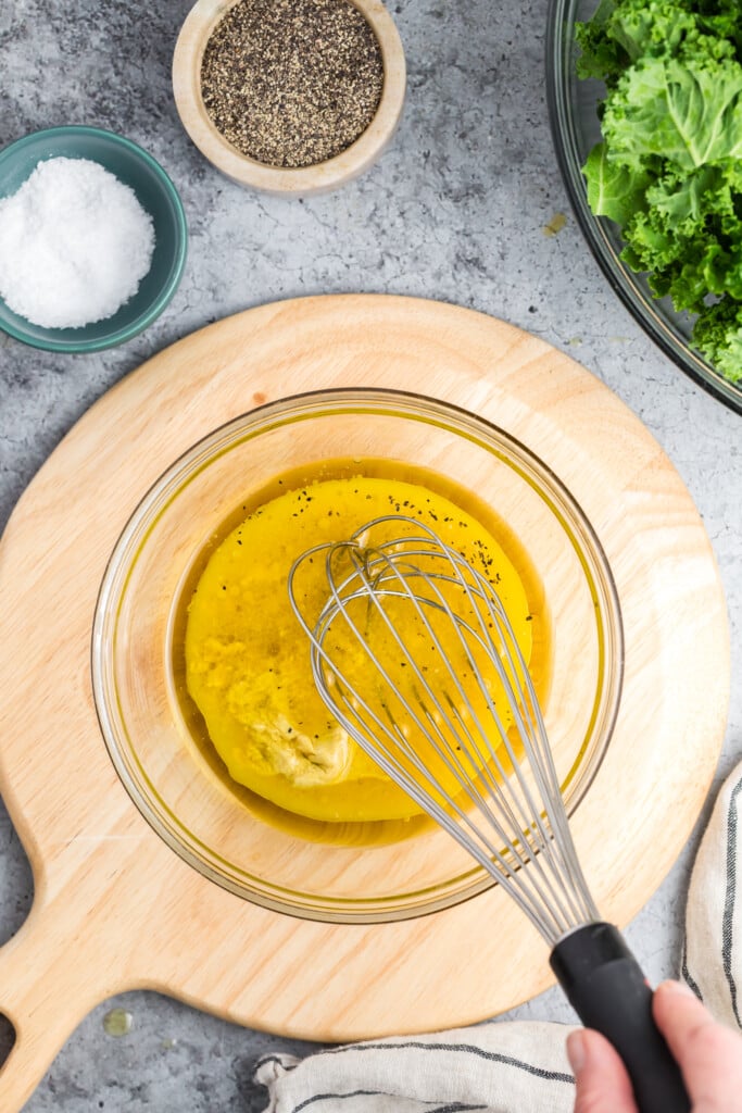 A hand uses a whisk to mix olive oil, mustard, and spices in a glass bowl on a wooden board. Nearby are bowls of salt, pepper, and fresh kale leaves.