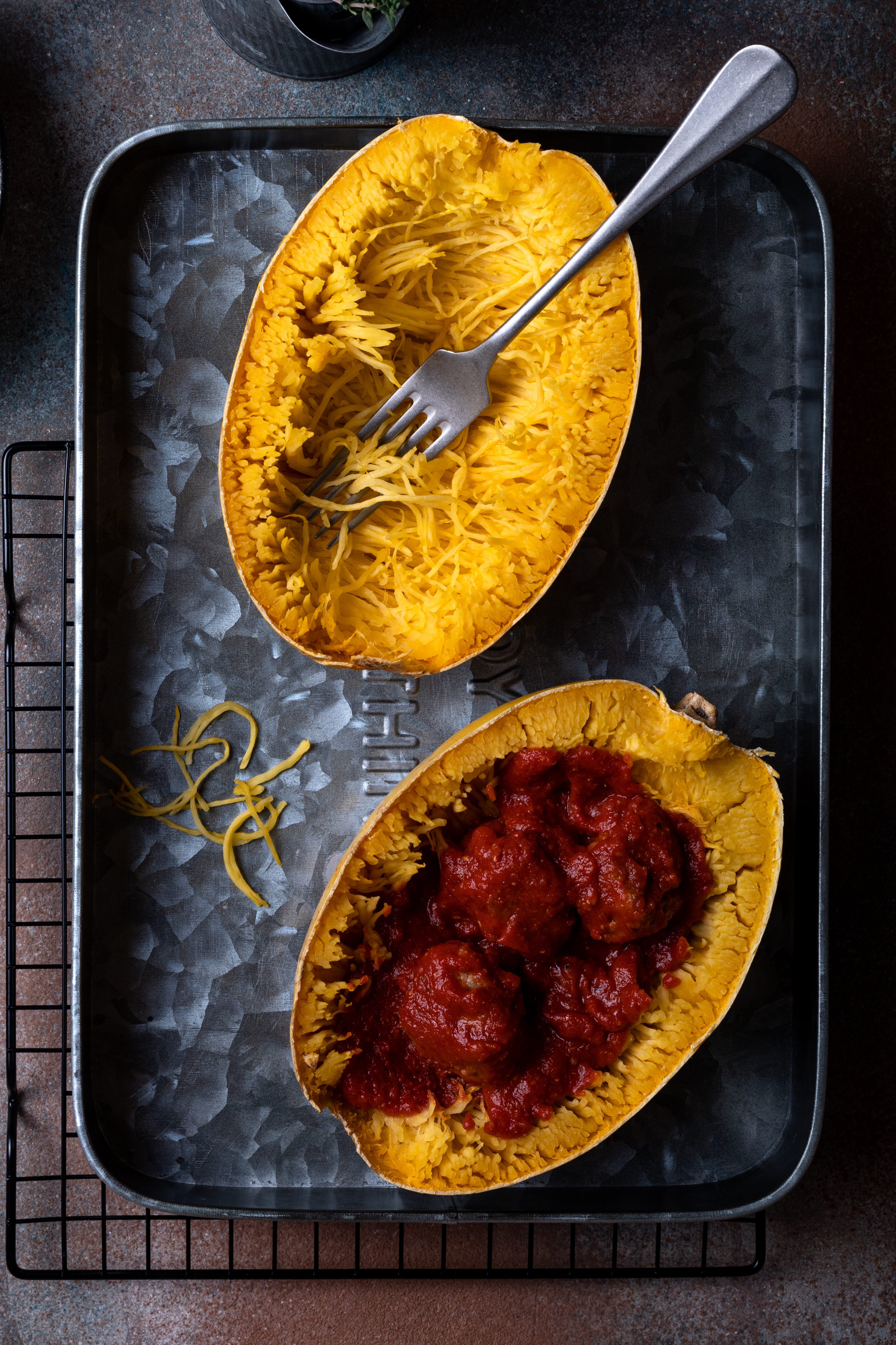 A baking tray with two halves of cooked spaghetti squash: one half is plain with a fork and some strands pulled up, the other half contains spaghetti squash topped with tomato sauce and meatballs.