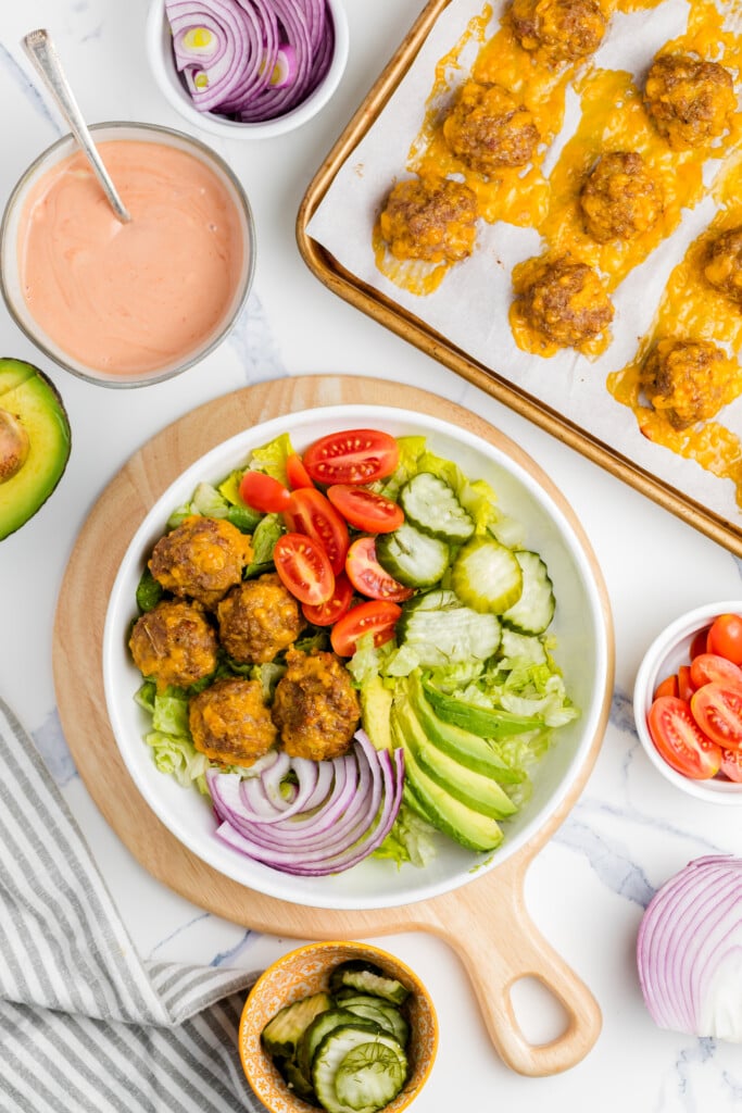A bowl with lettuce, cherry tomatoes, red onions, pickles, avocado slices, and small baked cheesy meatballs. Surrounding the bowl are a tray of meatballs, sauce, avocado, and bowls of tomatoes and pickles.