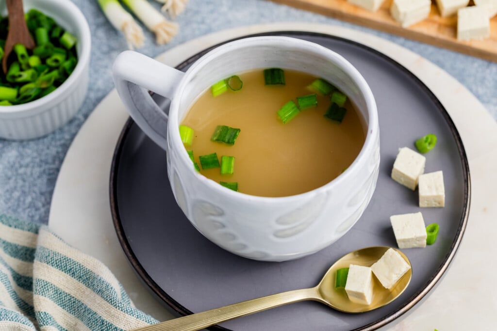 A white mug of miso soup garnished with green onions sits on a gray plate, with tofu cubes and a gold spoon beside it. A bowl of chopped green onions and a striped cloth are nearby.
