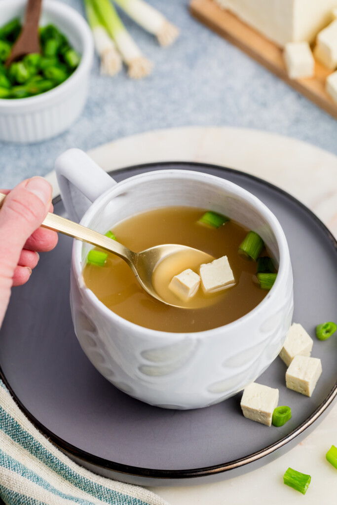 A hand holds a spoon above a white cup of clear soup with tofu cubes and chopped green onions. The cup sits on a gray plate, with extra tofu and green onions nearby on the table.