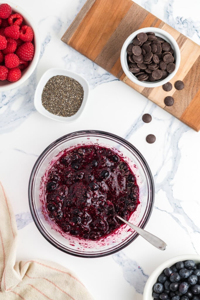 A bowl of berry chia jam with a spoon sits on a marble surface, surrounded by bowls of raspberries, blueberries, chia seeds, and chocolate chips, with a striped cloth partially visible.
