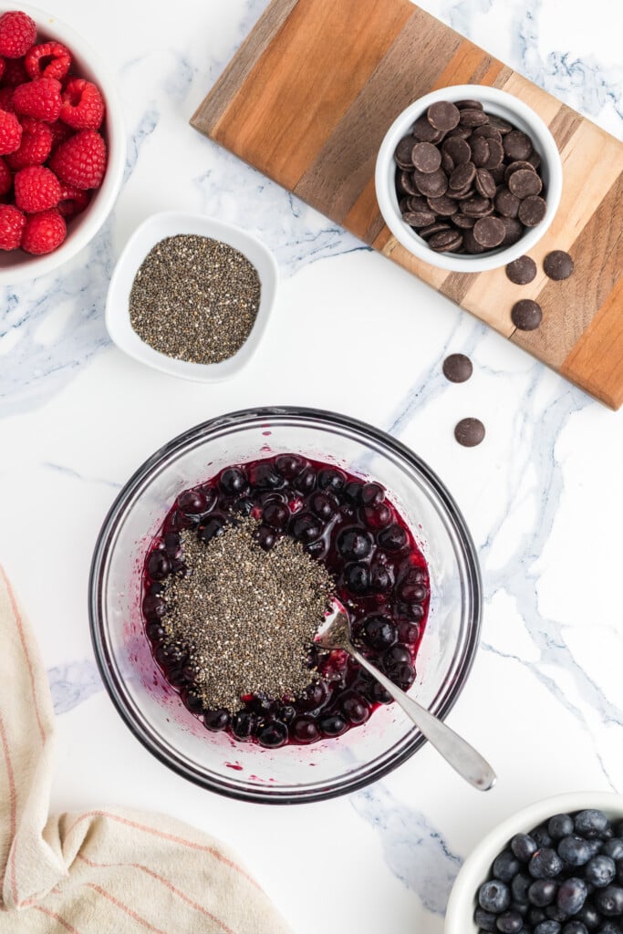 A bowl of blueberries mixed with chia seeds and a spoon on a marble countertop, surrounded by bowls of raspberries, chia seeds, blueberries, and chocolate chips on a wooden board.