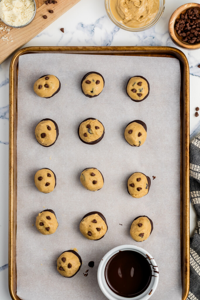 A baking sheet lined with parchment paper holds fifteen chocolate chip cookie dough balls, half-dipped in chocolate. Nearby are bowls of peanut butter, chocolate chips, flour, and a cup of melted chocolate.