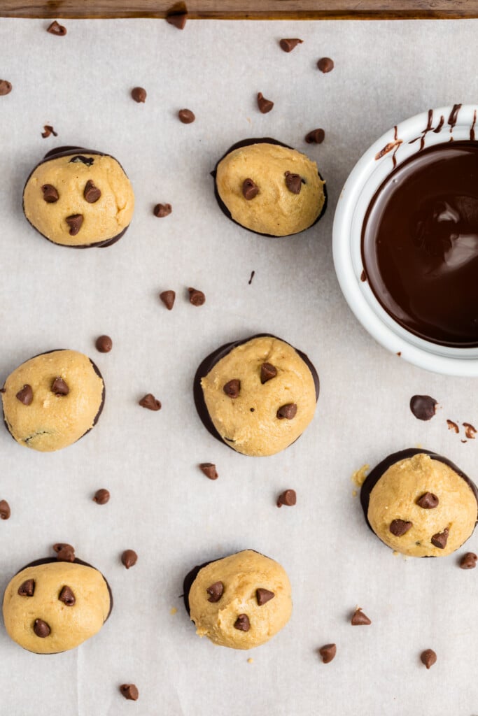 Overhead view of cookie dough balls with chocolate chips on parchment paper, some dipped in chocolate, scattered chocolate chips, and a white bowl of melted chocolate.