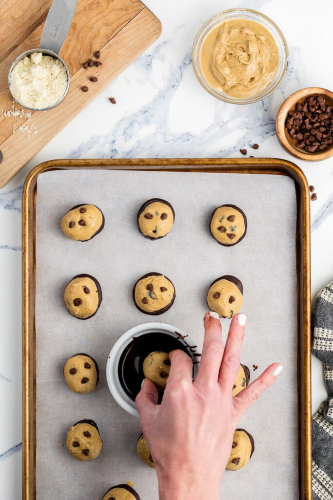 A hand dips a ball of cookie dough into melted chocolate above a baking tray lined with parchment paper, with more cookie dough balls, a bowl of peanut butter, chocolate chips, and flour on the side.