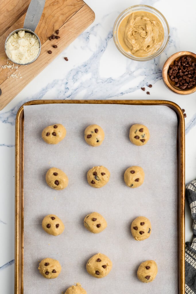A baking sheet lined with parchment paper holds unbaked chocolate chip cookie dough balls. Nearby are a bowl of peanut butter, a metal measuring cup with flour, a small bowl of chocolate chips, and a checkered towel.