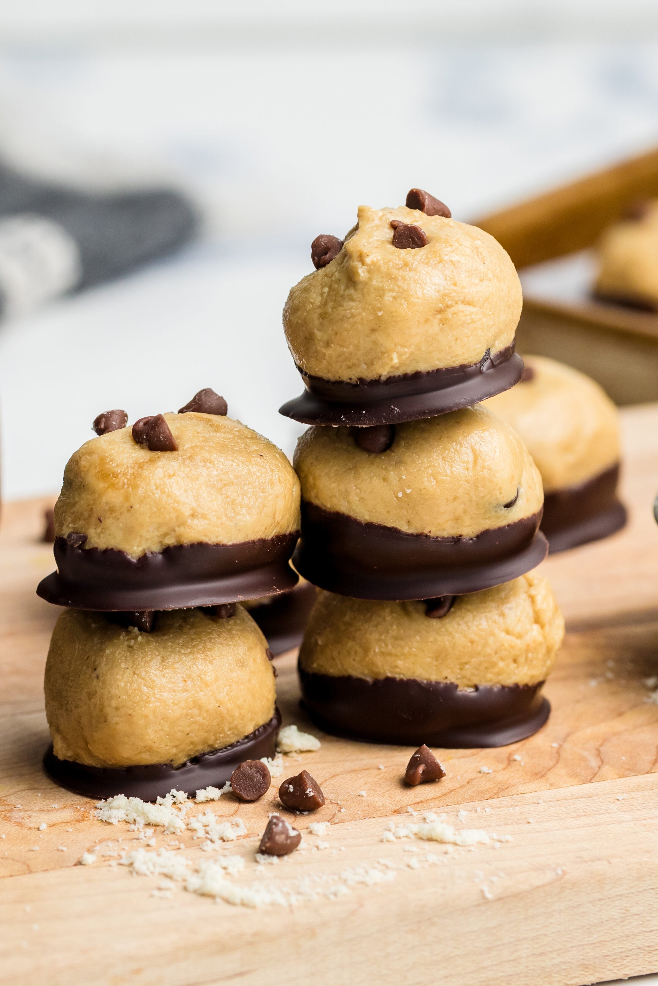Three stacks of cookie dough balls dipped in chocolate are on a wooden board, surrounded by chocolate chips, a measuring cup with flour, and more dough balls on a baking tray in the background.