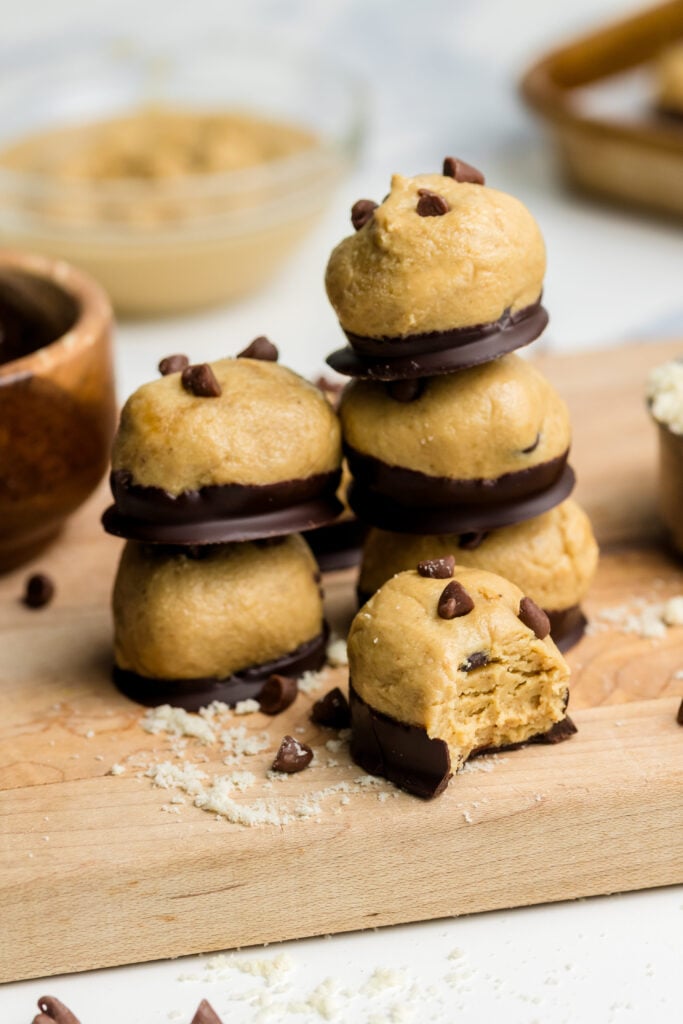 Three stacked cookie dough sandwiches with chocolate chips and chocolate filling are on a wooden board. A bitten piece, scattered chips, and baking ingredients are nearby; more sandwiches are on a tray in the background.