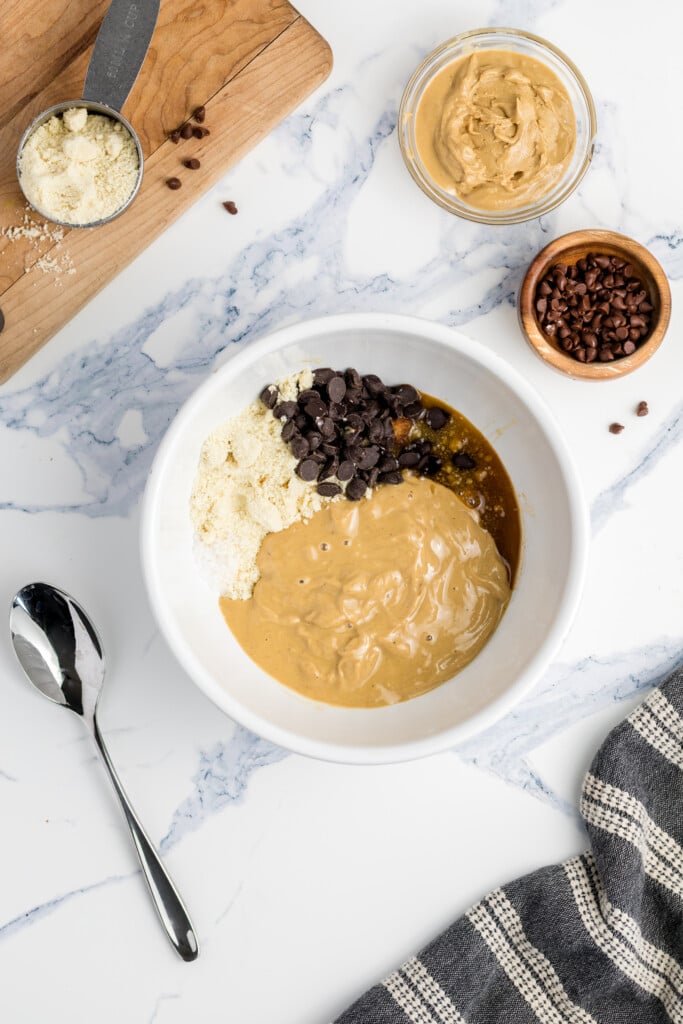 A white bowl on a marble countertop holds almond flour, chocolate chips, vanilla, and creamy peanut butter. Nearby are a spoon, a small bowl of chocolate chips, a bowl of peanut butter, a measuring cup, and a kitchen towel.
