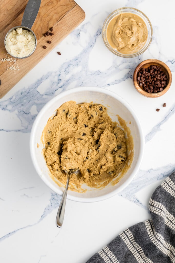 A white bowl filled with cookie dough and a spoon sits on a marble counter, surrounded by small bowls of peanut butter and chocolate chips, a measuring cup with flour, and a striped kitchen towel.