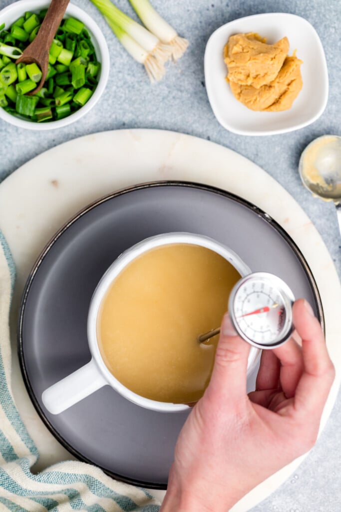 A hand holds a thermometer in a white pot of light brown broth, with chopped green onions, a bowl of miso paste, and a spoon nearby on a gray surface.