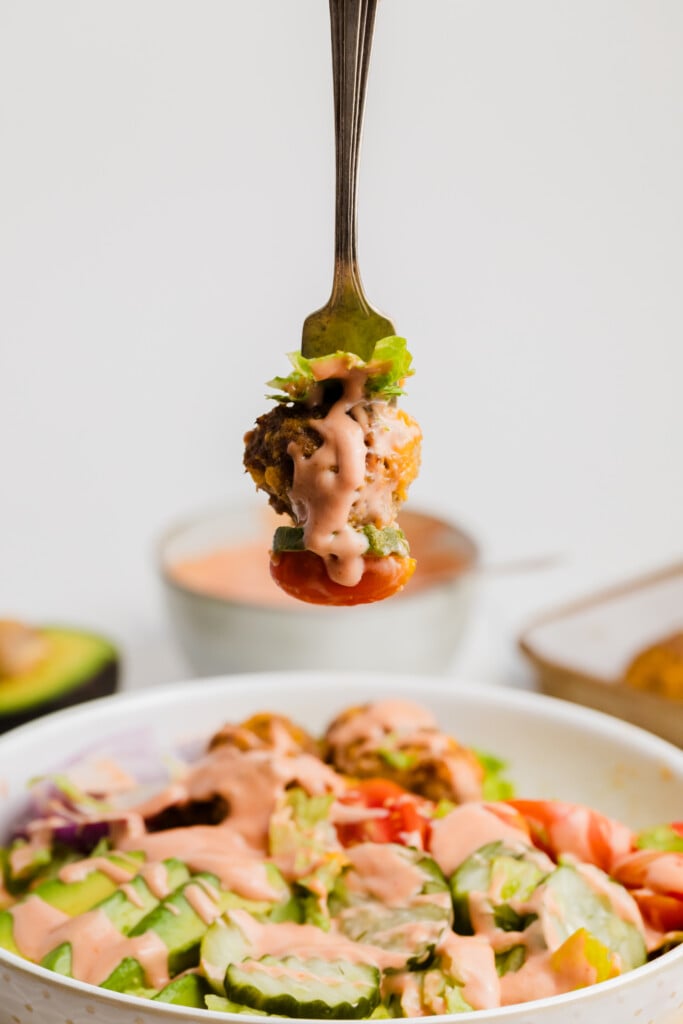 A fork holding a bite of salad with a veggie ball, lettuce, tomato, and creamy pink dressing hovers above a salad bowl filled with cucumbers, tomatoes, and more dressing. Avocado and sauce are blurred in the background.
