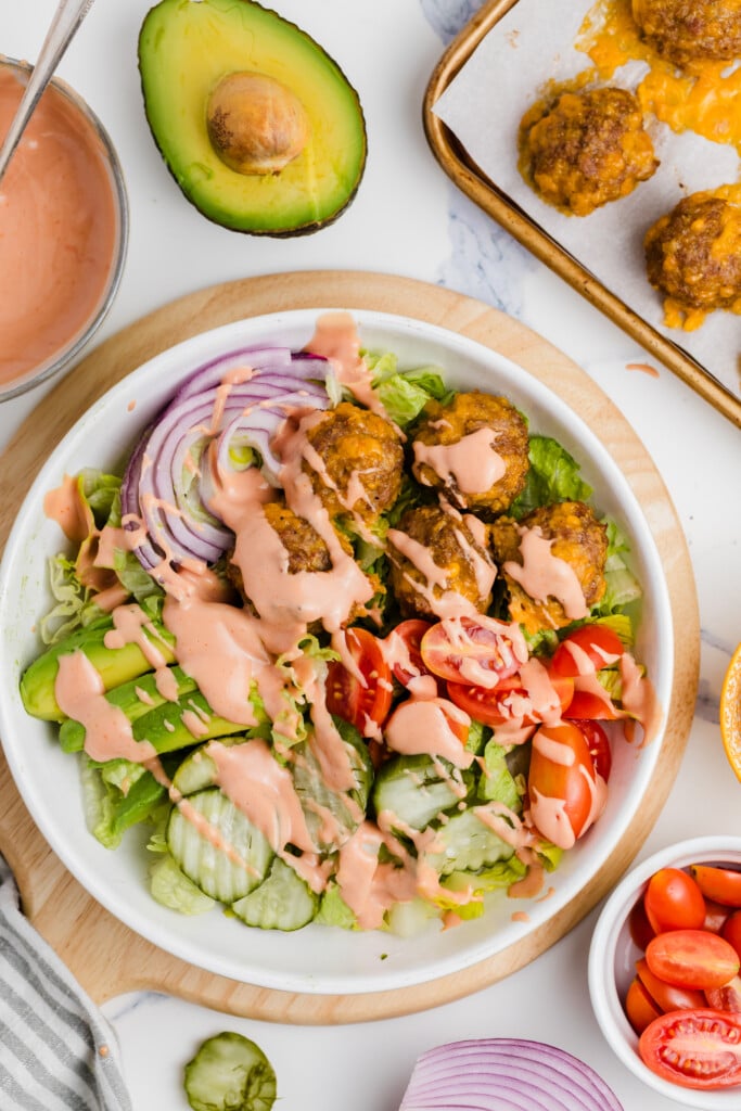 A bowl of salad with lettuce, cucumbers, cherry tomatoes, red onion, avocado slices, and meatballs, topped with pink dressing. Sliced avocado, sauce, and a tray of meatballs are nearby.