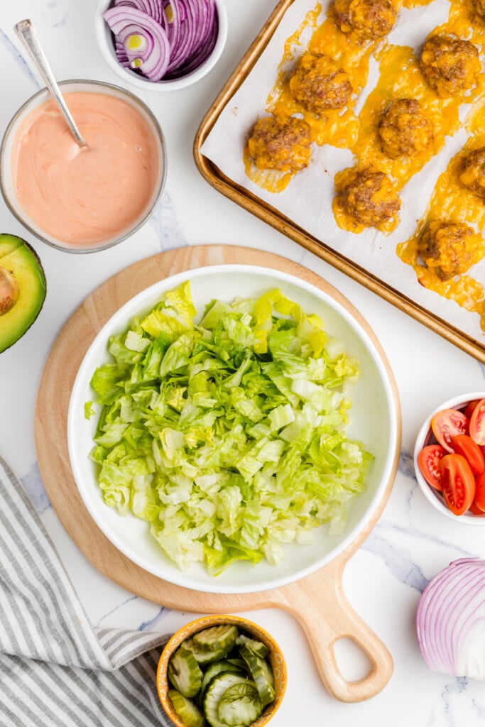 A bowl of chopped lettuce on a wooden board, surrounded by sliced pickles, red onion, halved cherry tomatoes, half an avocado, a bowl of dressing, and a tray of cheesy baked meatballs on parchment paper.