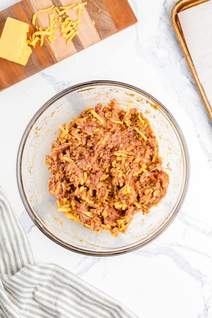 A glass bowl contains a mixture of ground meat and shredded cheese. Nearby, a wooden board holds a block of cheddar and scattered cheese, while a baking sheet with parchment paper sits to the side on a marble countertop.