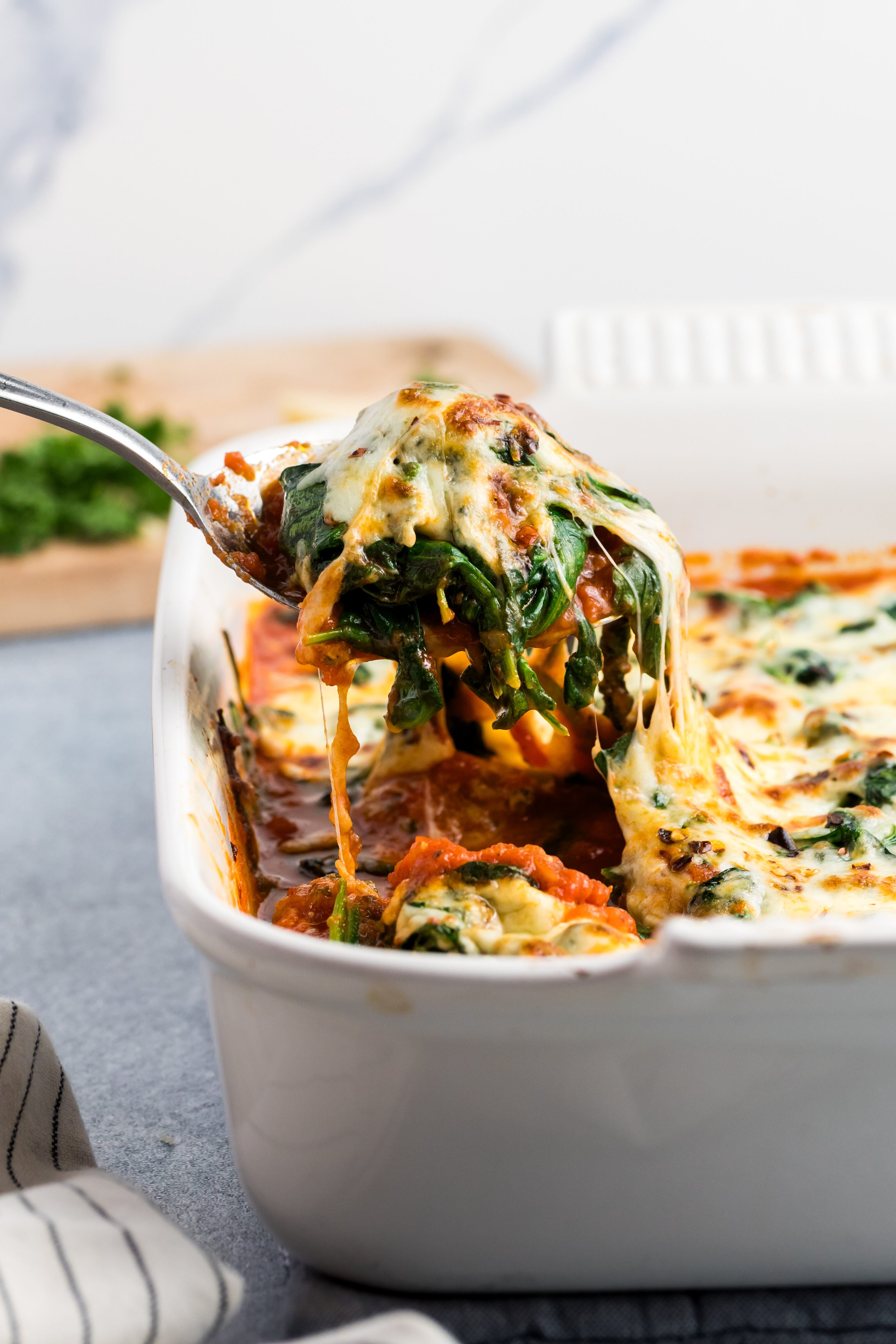 A spoon lifts a serving of cheesy baked casserole with spinach and vegetables from a white ceramic dish, with melted cheese stretching. The background features a light marble wall and a wooden board with chopped herbs.
