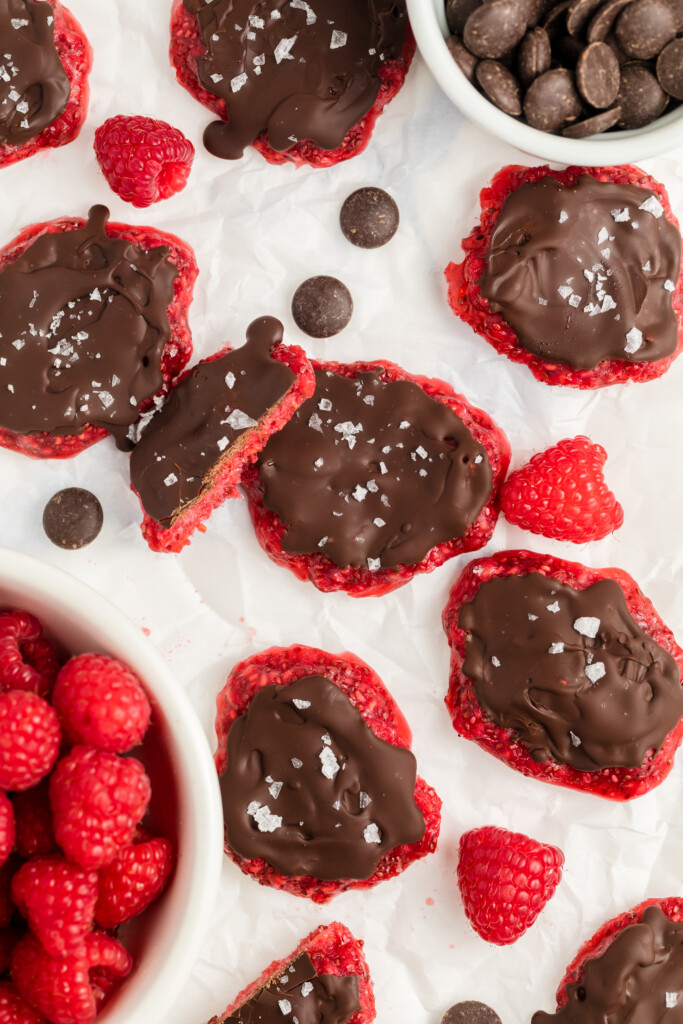 Red cookies topped with melted chocolate and flaky sea salt are arranged on parchment paper, surrounded by fresh raspberries, chocolate wafers, and scattered chocolate pieces. One cookie is partially eaten.