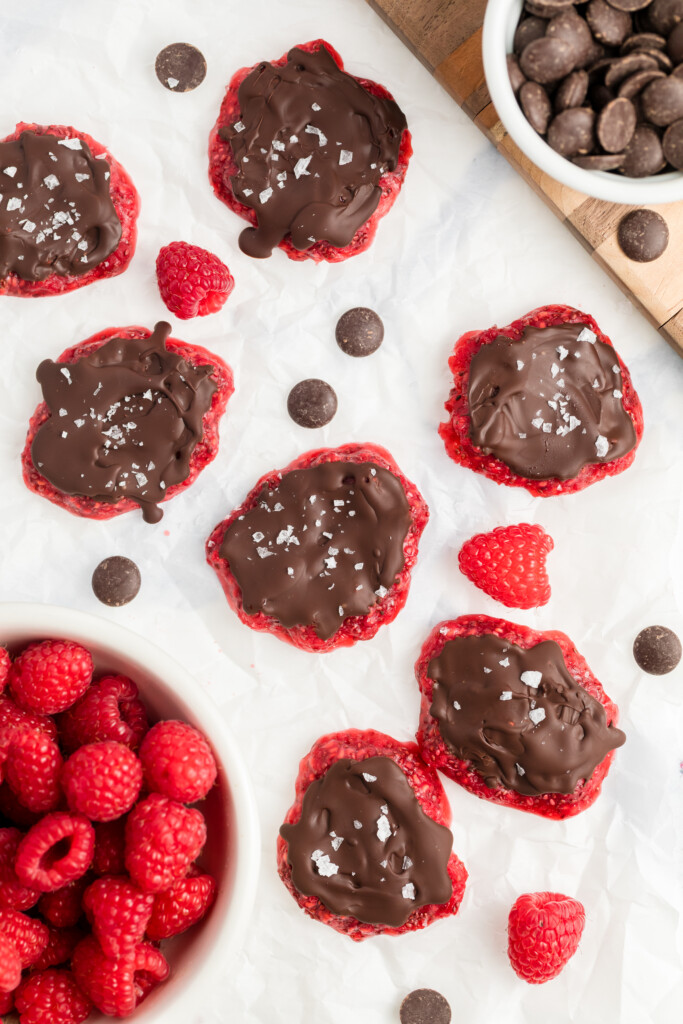 Chocolate-covered raspberry clusters topped with sea salt are arranged on parchment paper, surrounded by fresh raspberries and chocolate pieces, with bowls of raspberries and chocolate nearby.
