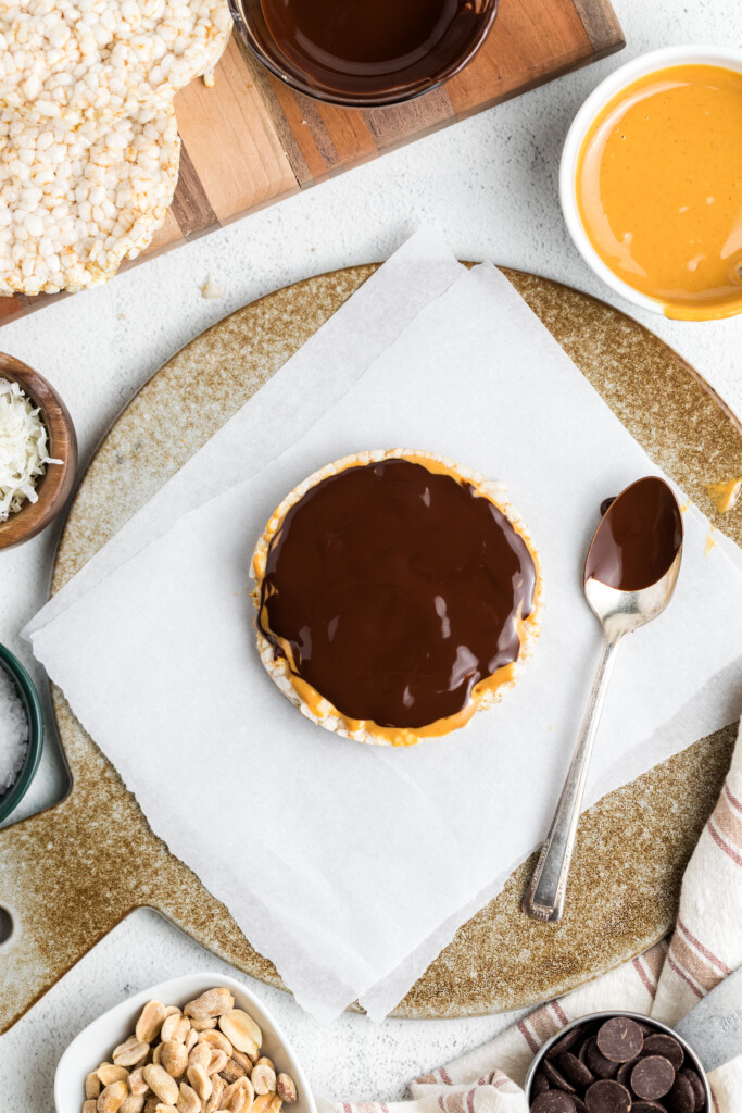 A Chocolate Peanut Butter Rice Cake Treat sits on parchment paper, surrounded by a spoon, peanuts, shredded coconut, chocolate chips, and a bowl of peanut butter.